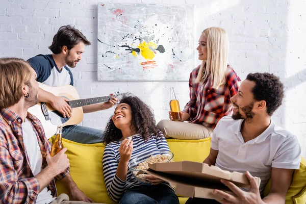 Jovem tocando guitarra acústica para amigos multiculturais alegres durante a festa — Fotografia de Stock