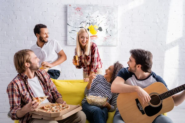 Молодой человек играет на акустической гитаре рядом со счастливыми многоэтническими друзьями во время вечеринки — стоковое фото