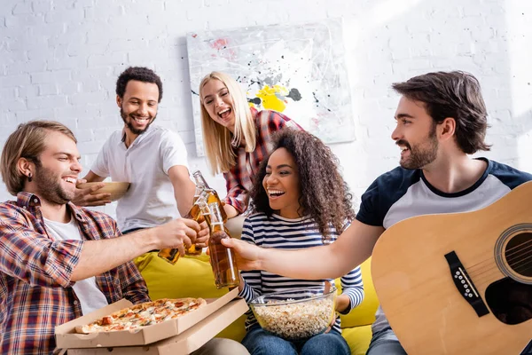 Joven sosteniendo la guitarra acústica mientras tintinea botellas de cerveza con amigos multiétnicos - foto de stock