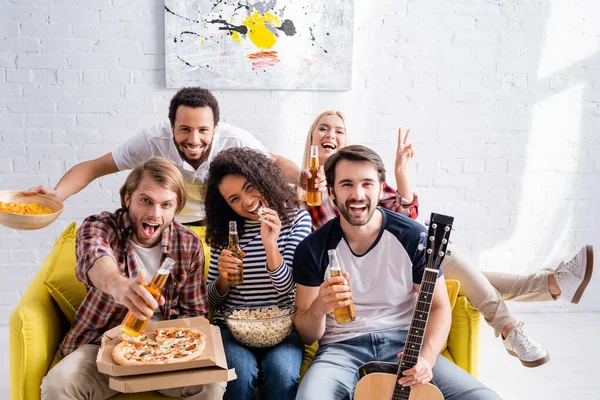 Cheerful woman showing peace gesture and man holding acoustic guitar near excited multicultural friends with beer — Stock Photo