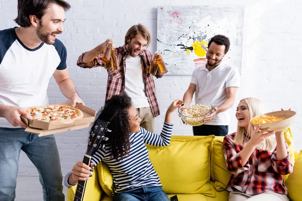 Mulher afro-americana com guitarra levando pipocas perto de amigos multiculturais excitados segurando cerveja e lanches — Fotografia de Stock