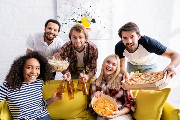 Веселый мультикультурный друг с пивом, пиццей и закусками, смотрящий в камеру во время вечеринки — стоковое фото