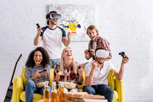 KYIV, UCRANIA - 19 de octubre de 2020: hombres jóvenes con auriculares vr que muestran un gesto de victoria cerca de amigos multiétnicos emocionados jugando videojuegos - foto de stock