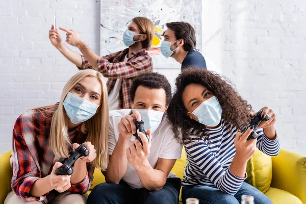 KYIV, UKRAINE - OCTOBER 19, 2020: positive multicultural friends in medical masks playing video game near man pointing at smartphone on blurred background — Stock Photo