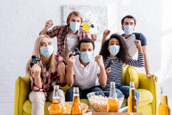 KYIV, UKRAINE - OCTOBER 19, 2020: excited multiethnic friends in medical masks showing win gesture while playing video game near beer on blurred foreground — Stock Photo
