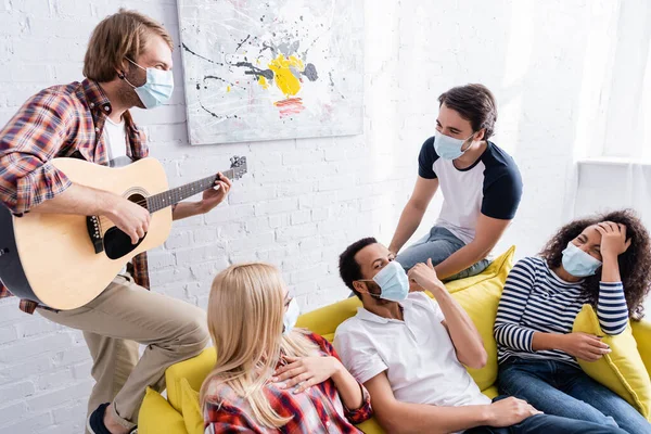 Young man playing acoustic guitar to multiethnic friends in medical masks during party — Stock Photo
