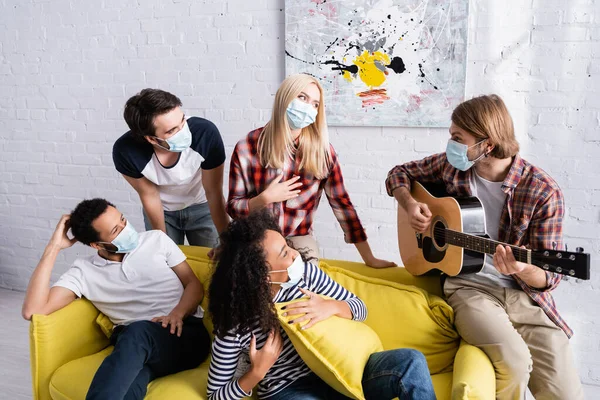 Young man playing acoustic guitar to multicultural friends in medical masks sitting on sofa during party — Stock Photo