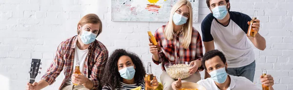 Multicultural friends in medical masks holding bottles of beer and looking at camera, banner — Stock Photo