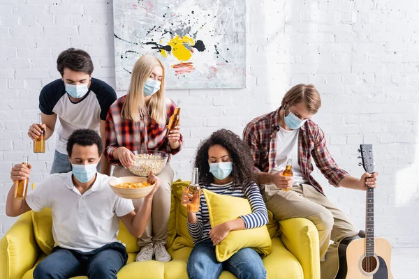 Multicultural friends in medical masks holding beer and snacks with closed eyes during party — Stock Photo