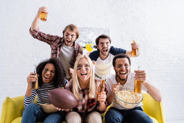 Excited multiethnic friends with painted faces holding beer and screaming while watching rugby championship — Stock Photo