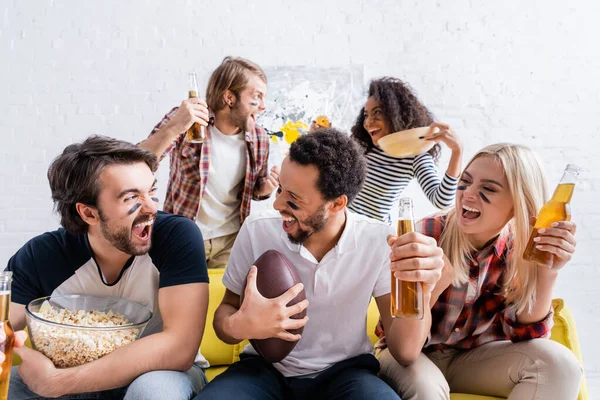 Alegres aficionados multiétnicos al rugby con caras pintadas sosteniendo la cerveza mientras observan la competencia en casa - foto de stock