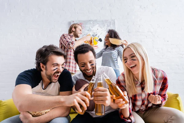 Hombre emocionado gritando mientras tintinea botellas de cerveza con amigos multiculturales con caras pintadas - foto de stock