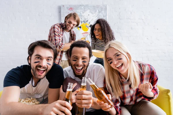 Cheerful multicultural rugby fans with painted faces clinking bottles of beer near friends on blurred background — Stock Photo