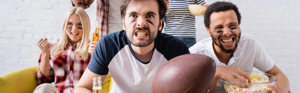 Giovane uomo che tiene palla da rugby e smorfia mentre guarda il campionato vicino agli amici multietnici, striscione — Foto stock