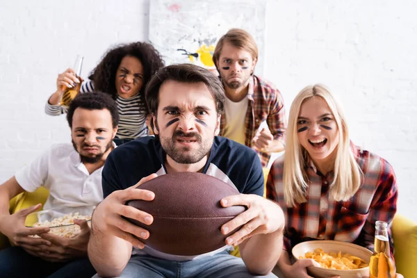 Joven sosteniendo pelota de rugby cerca de amigos multiétnicos con caras pintadas viendo el campeonato sobre fondo borroso - foto de stock