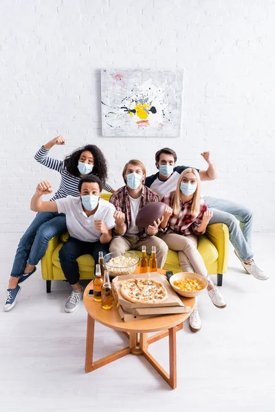 High angle view of multiethnic rugby fans in medical masks showing win gesture while watching championship at home — Stock Photo