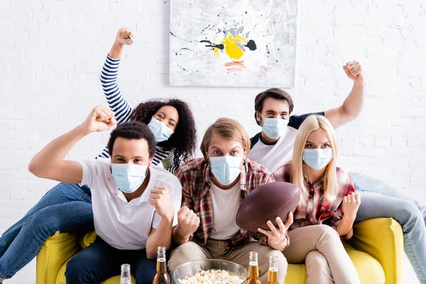 Excited multicultural friends in medical masks showing win gesture while watching rugby championship — Stock Photo