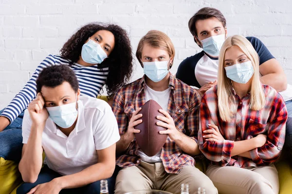 Upset man holding rugby ball near sad multiethnic friends in medical masks watching competition at home — Stock Photo