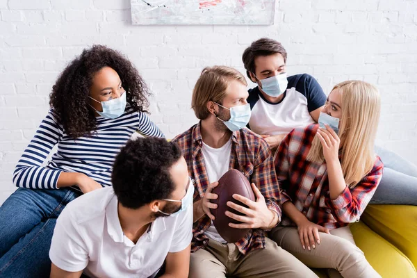 Young man holding rugby ball while talking with multicultural friends in medical masks — Stock Photo