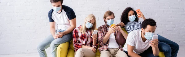 Багатоетнічні друзі в медичних масках обміну повідомленнями на смартфонах, сидячи на дивані, банер — стокове фото
