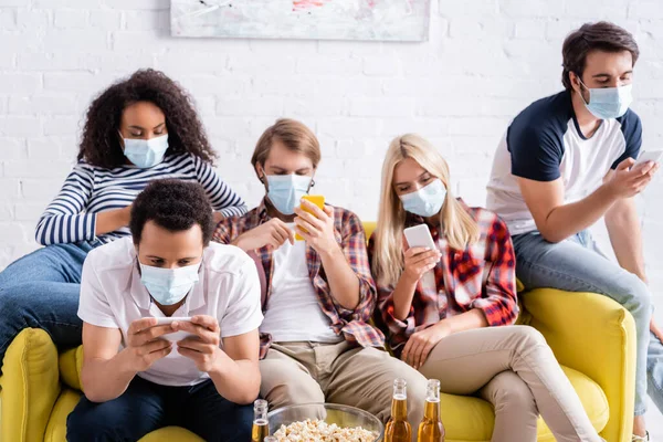 Молоді багатоетнічні друзі в медичних масках обміну повідомленнями на смартфонах, сидячи на дивані — стокове фото