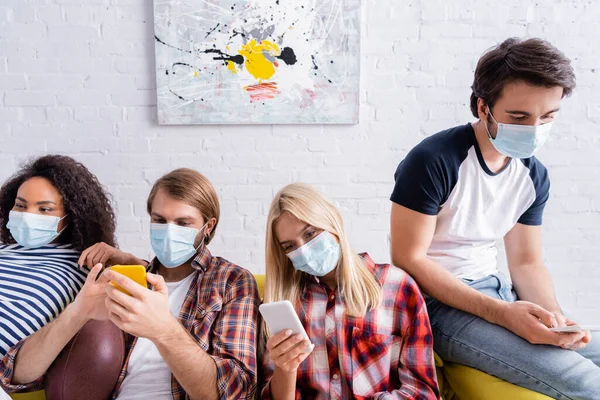 Багатоетнічні друзі в медичних масках обміну повідомленнями на мобільних телефонах, сидячи на дивані — стокове фото