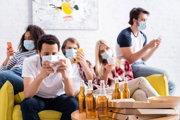 Amici multietnici in maschere mediche messaggistica sui telefoni cellulari vicino a birra e pizza, sfondo sfocato — Foto stock