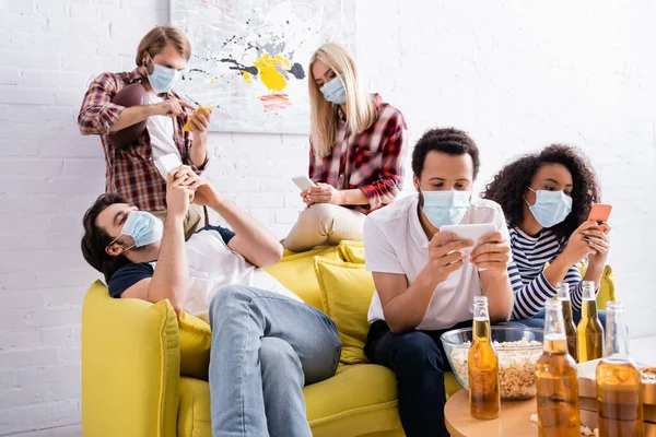 Multicultural friends in medical masks chatting on smartphones while sitting on sofa during party — Stock Photo