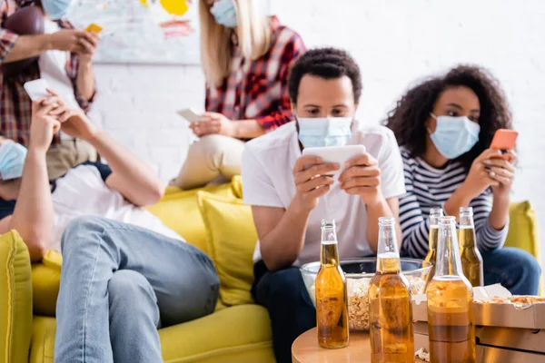 Bottles of beer and bowl of popcorn near multiethnic friends in medical masks messaging on mobile phones on blurred background — Stock Photo