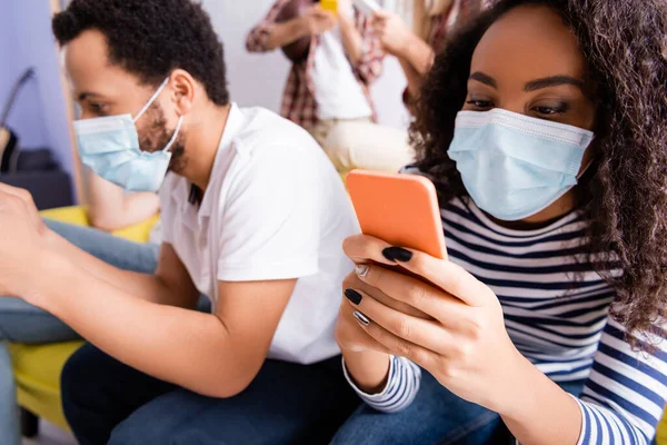 Donna afroamericana in maschera medica messaggistica sul telefono cellulare vicino amici multiculturali su sfondo sfocato — Foto stock
