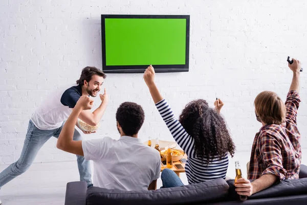 Aufgeregter Mann zeigt auf LCD-Fernseher an Wand in der Nähe multikultureller Freunde, die Sportwettkämpfe verfolgen — Stockfoto