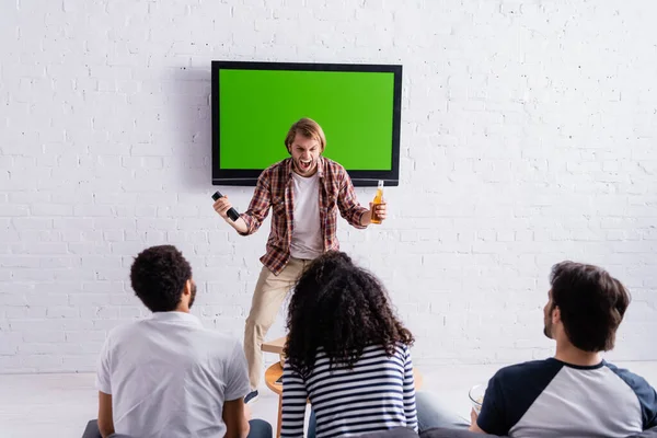 Aufgeregter Mann schreit und zeigt Siegergeste in der Nähe des LCD-Fernsehers, während multikulturelle Freunde Sportwettkämpfe verfolgen — Stockfoto