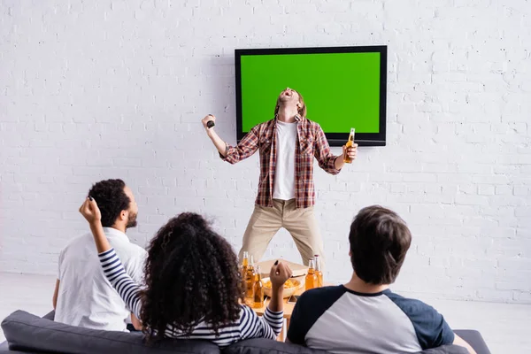 Hombre emocionado gritando y mostrando el gesto de ganar cerca de lcd tv y aficionados a los deportes multiculturales - foto de stock