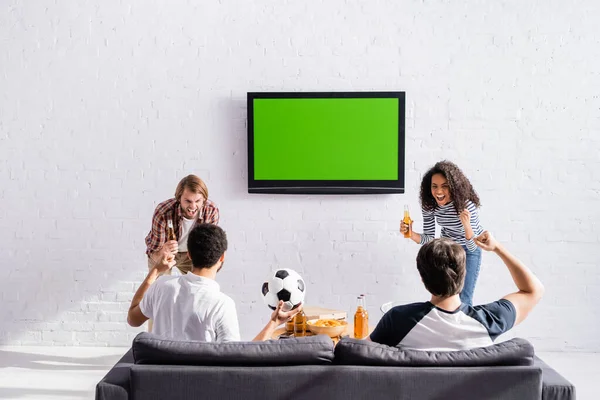 Excité amis multiethniques montrant geste de victoire tout en tenant de la bière près de lcd tv sur le mur — Photo de stock