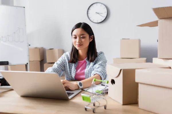 Sorridente asiatico volontario digitando sul computer portatile, mentre seduto alla scrivania con scatole di cartone e carrello della spesa in ufficio di beneficenza — Foto stock