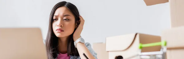 Nachdenkliche asiatische Frau blickt auf Laptop in der Nähe von Kartons auf verschwommenem Vordergrund, Banner — Stockfoto