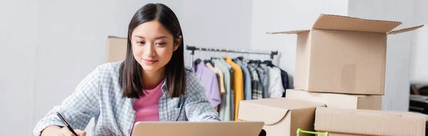 Sonriente asiático voluntario con pluma mirando portátil cerca de cajas de cartón en el centro de caridad, pancarta - foto de stock