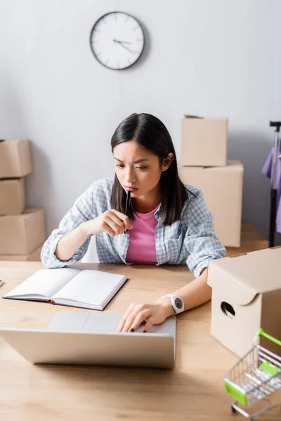 Nachdenklicher asiatischer Freiwilliger mit Stift blickt auf Laptop, während er am Schreibtisch mit Notizbuch, Karton und Einkaufswagen sitzt — Stockfoto