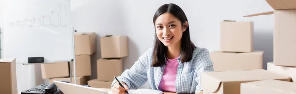 Happy asian volunteer looking at camera near carton boxes in charity center, banner — Stock Photo