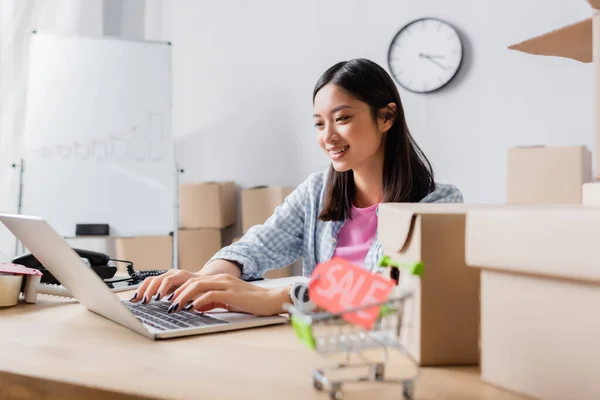 Happy asian volunteer typing on laptop while sitting at desk with carton boxes in charity center on blurred foreground — Stock Photo