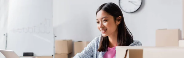 Happy asian volunteer looking at laptop near carton boxes in charity center on blurred foreground, banner — Stock Photo
