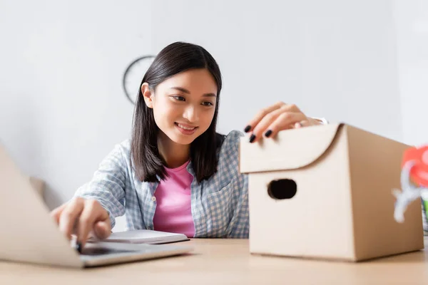 Feliz asiático voluntário olhando caixa de papelão enquanto sentado na mesa no centro de caridade em primeiro plano borrado — Fotografia de Stock