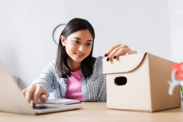 Glücklicher asiatischer Freiwilliger blickt auf Karton, während er am Schreibtisch im Charity Center auf verschwommenem Vordergrund sitzt — Stockfoto