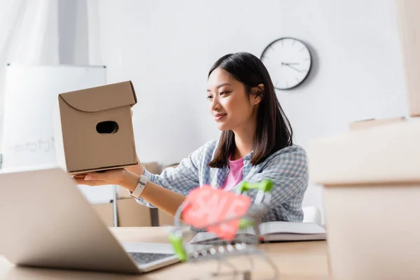 Smiling asian volunteer holding and looking at carton box near desk in charity center on blurred foreground — Stock Photo