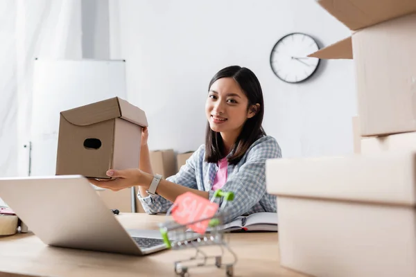 Souriant asiatique bénévole avec boîte en carton regardant la caméra près du bureau avec ordinateur portable et panier flou au premier plan — Photo de stock