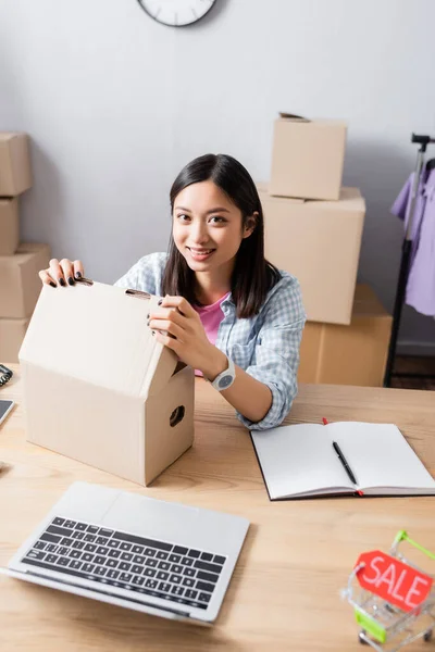 Smiling asian volunteer looking at camera while opening carton box at desk with notebook and laptop in charity center — Stock Photo