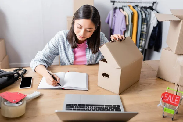 Asian volunteer opening carton box and writing in notebook at desk with digital devices in charity center — Stock Photo