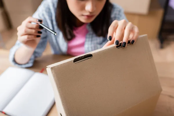 Cropped view of woman pointing with pen while opening carton box on blurred background — Stock Photo