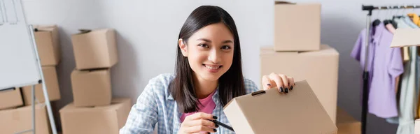 Happy asian volunteer looking at camera while opening carton box in charity center on blurred background, banner — Stock Photo