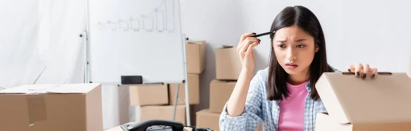 Afraid asian volunteer with pen near opened carton box in charity office, banner — Stock Photo